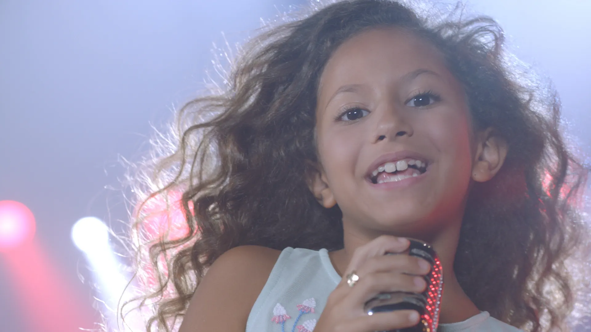 A smiling young singer with curly hair holding a microphone under pink stage light