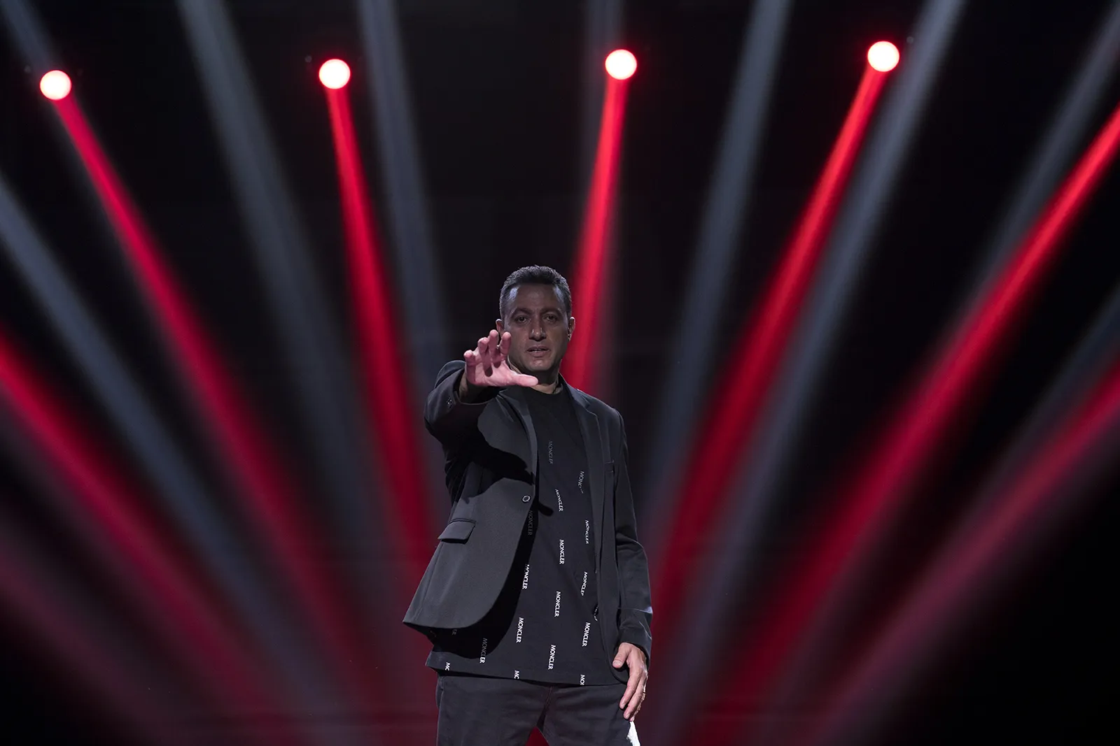 Waleed Nassif on a dark stage under red light beams, framing the shot with his hand