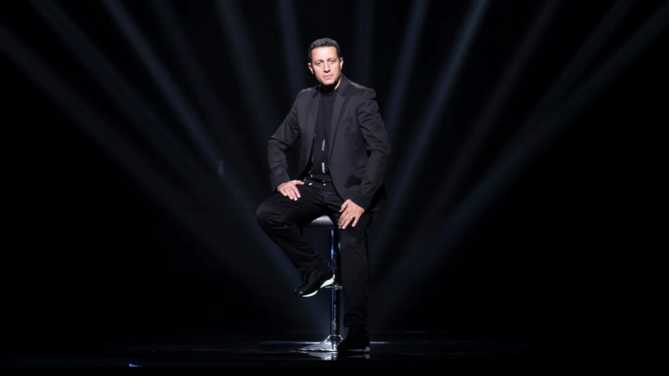 Waleed Nassif seated on a stool on a dark stage under fanned white light beams