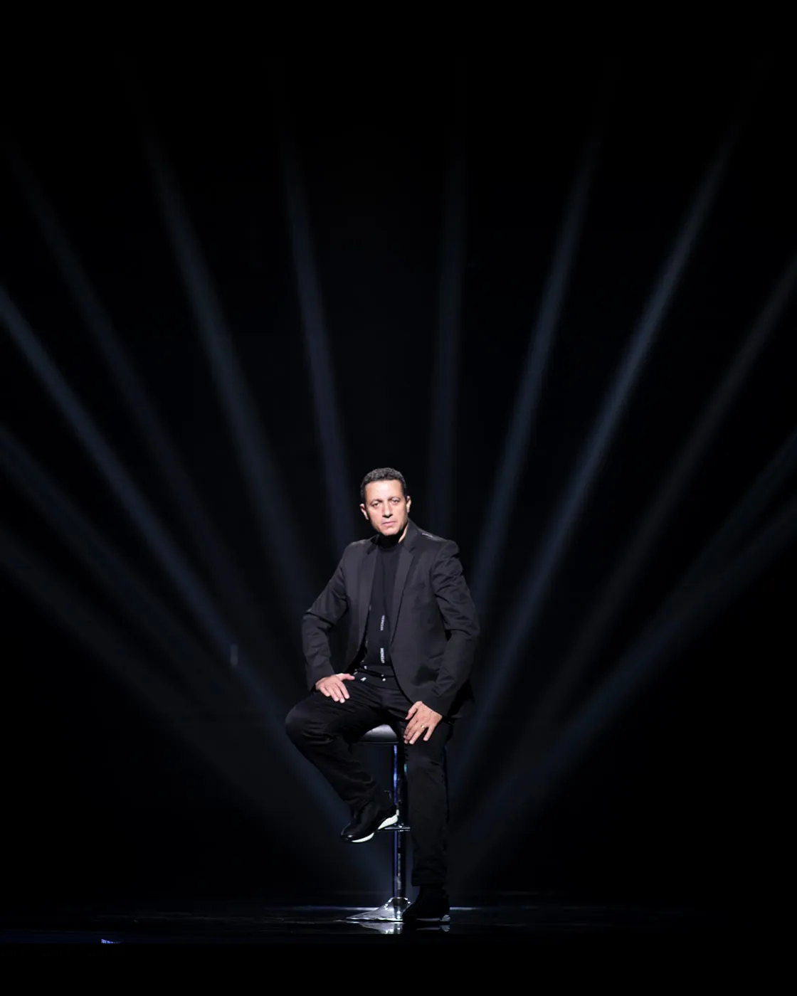 Waleed Nassif seated on a stool on a dark stage under fanned white light beams